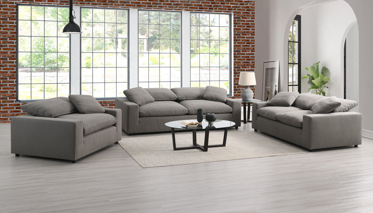 A modern living room with a Peyton Classic Sofa around a round coffee table. Large windows, a brick wall accent, light wood flooring, and a potted plant by the archway add to the décor, ideal for contemporary spaces.