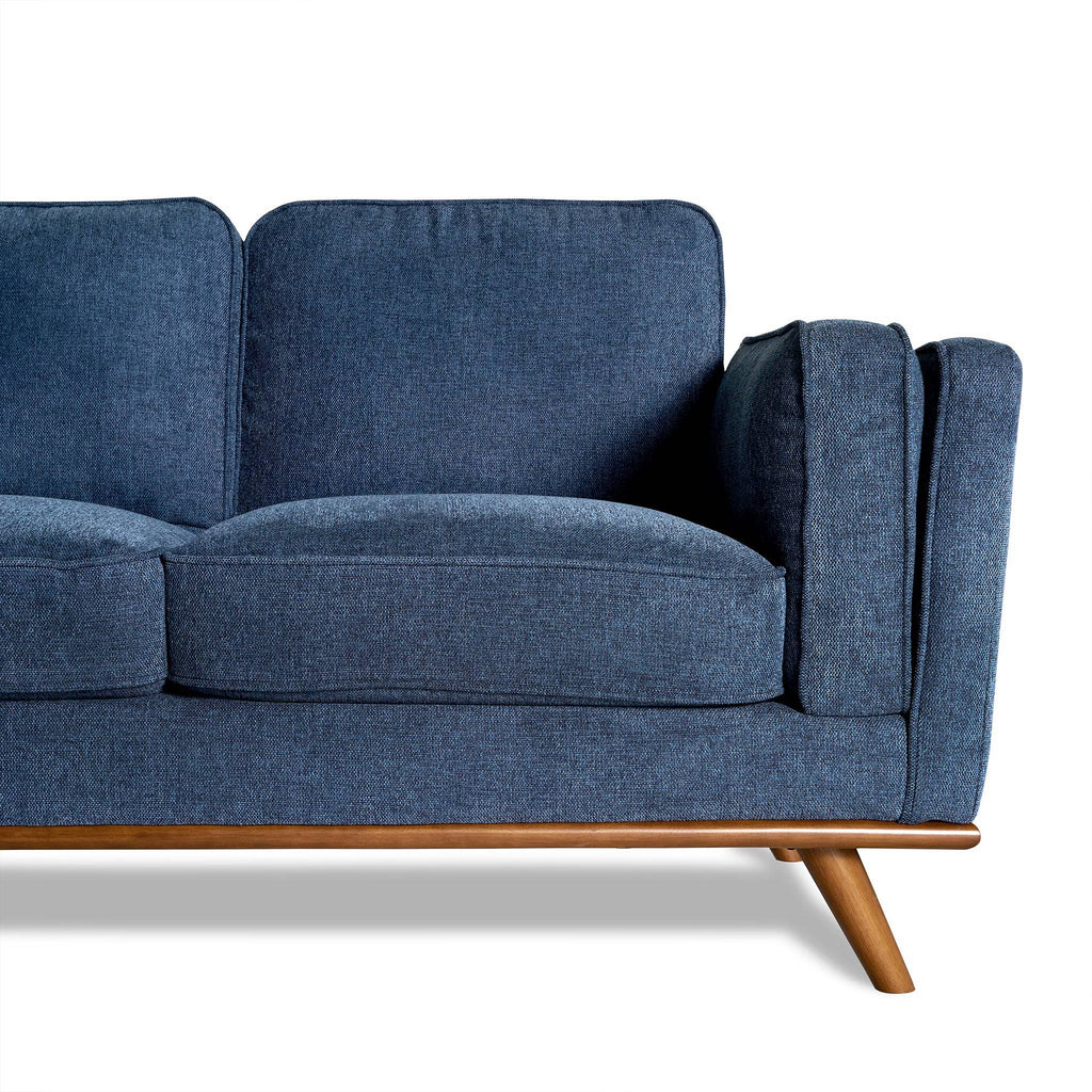 The Maddox Sectional, with its blue fabric upholstery and mid-century modern style, features wooden legs and armrests. Set against a white background, its plush cushions bring elegance to any living room. The focus is on the right half of this minimalist design.