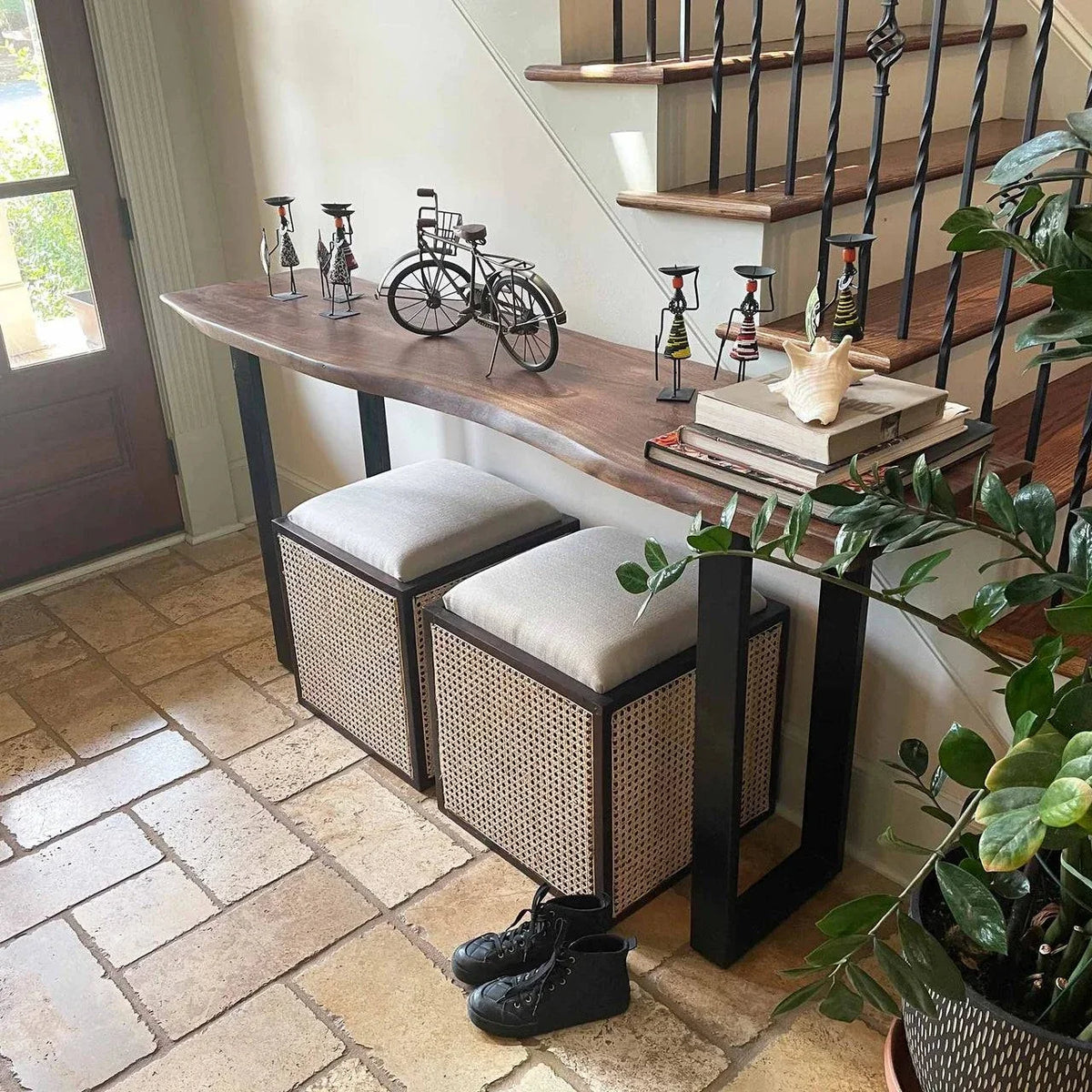 A hallway showcases the Live Edge Console adorned with artistic bicycle sculptures, books, and a shell. Below sit two uniquely styled storage benches. Nearby are black shoes and a plant on eco-friendly tile flooring, with stairs featuring a black railing in the background.