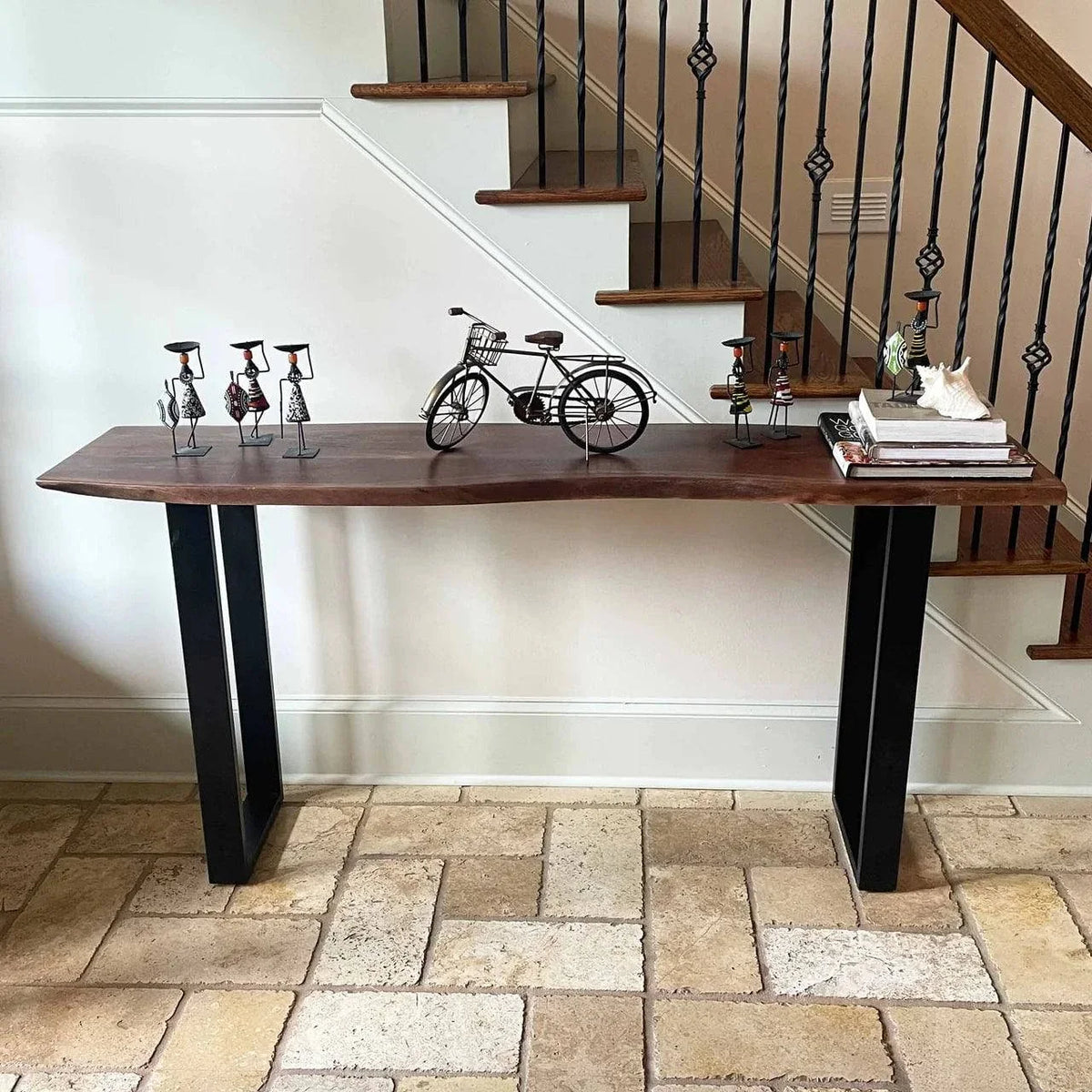 The Live Edge Console, made from acacia wood with black metal legs, features metal bicycle sculptures and figures. On a stone-tiled floor near a staircase with black wrought iron railings, its adorned with books and an eco-friendly white decor item.