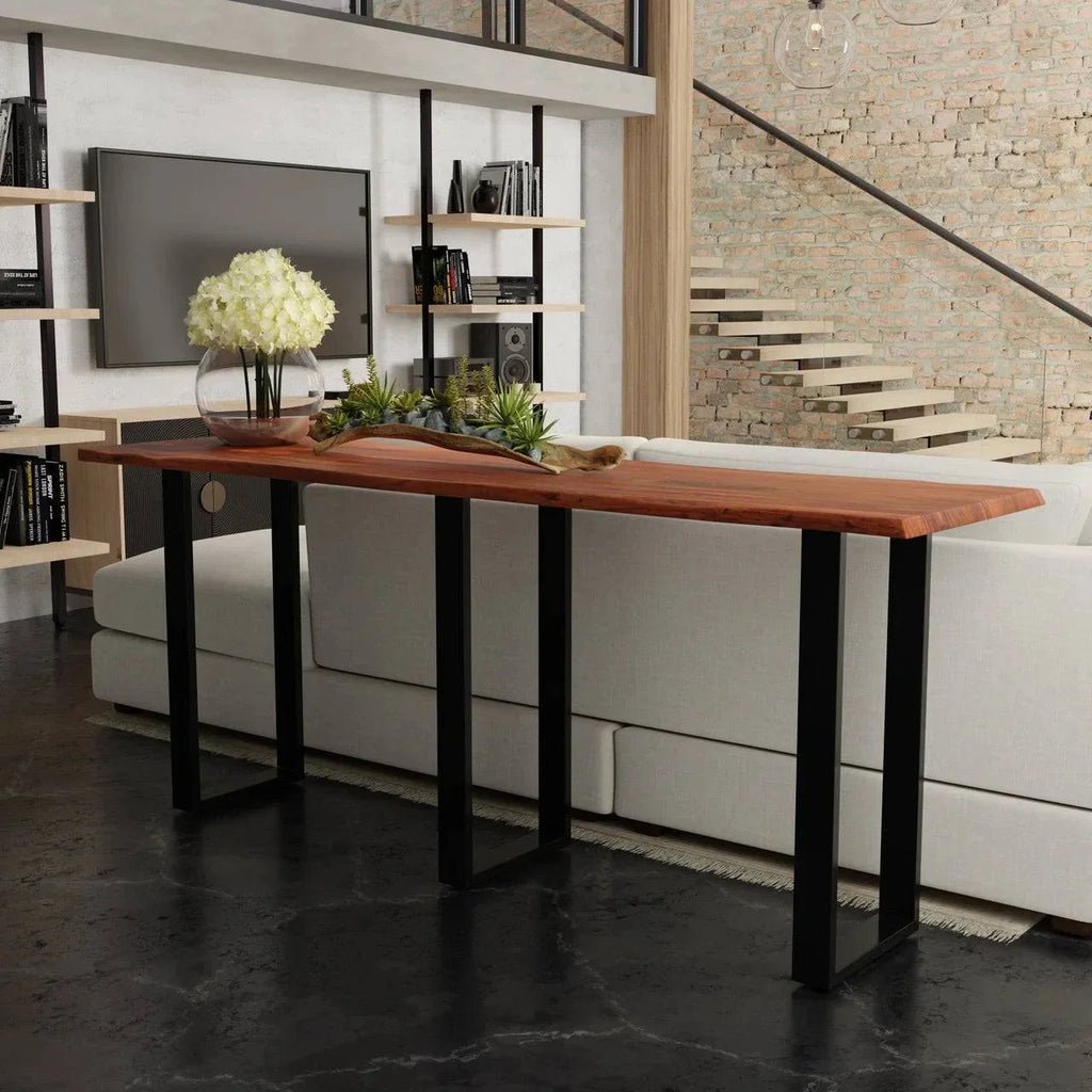 A stylish living room showcases the Live Edge Console, featuring a live edge acacia wood top and black metal legs, adorned with a glass vase of white flowers and a decor item. In the background, theres a white couch, exposed brick wall, bookshelves, and staircase.