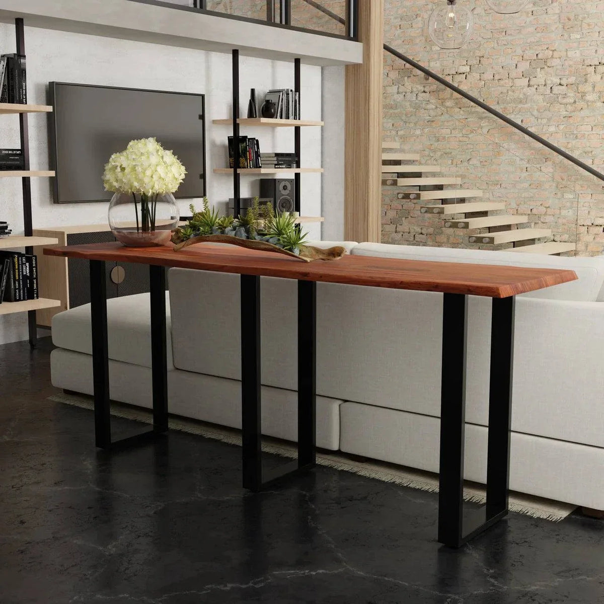 A stylish living room showcases the Live Edge Console, featuring a live edge acacia wood top and black metal legs, adorned with a glass vase of white flowers and a decor item. In the background, theres a white couch, exposed brick wall, bookshelves, and staircase.